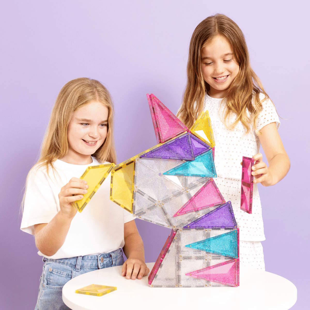 Two children playing with Connetix magnetic tiles building a colorful geometric unicorn against a purple background