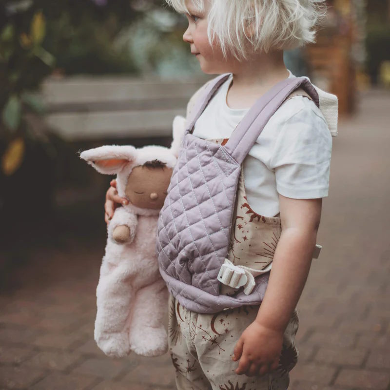 Child holding an Olli Ella Cozy Dinkum Bunny Pip in pale pink and wearing a lavender Doll Carrier