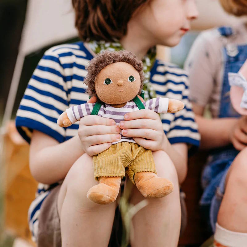 Child holding an Olli Ella Dress Me Dinky Doll with a striped shirt and suspenders, sitting outdoors.