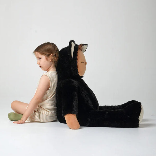Child in a white dress sitting next to a very large black cat plush toy that is bigger than her