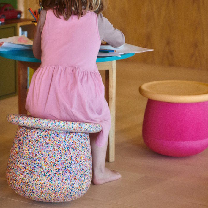 Child sitting on a colorful stool at a table with a pink chair nearby.
